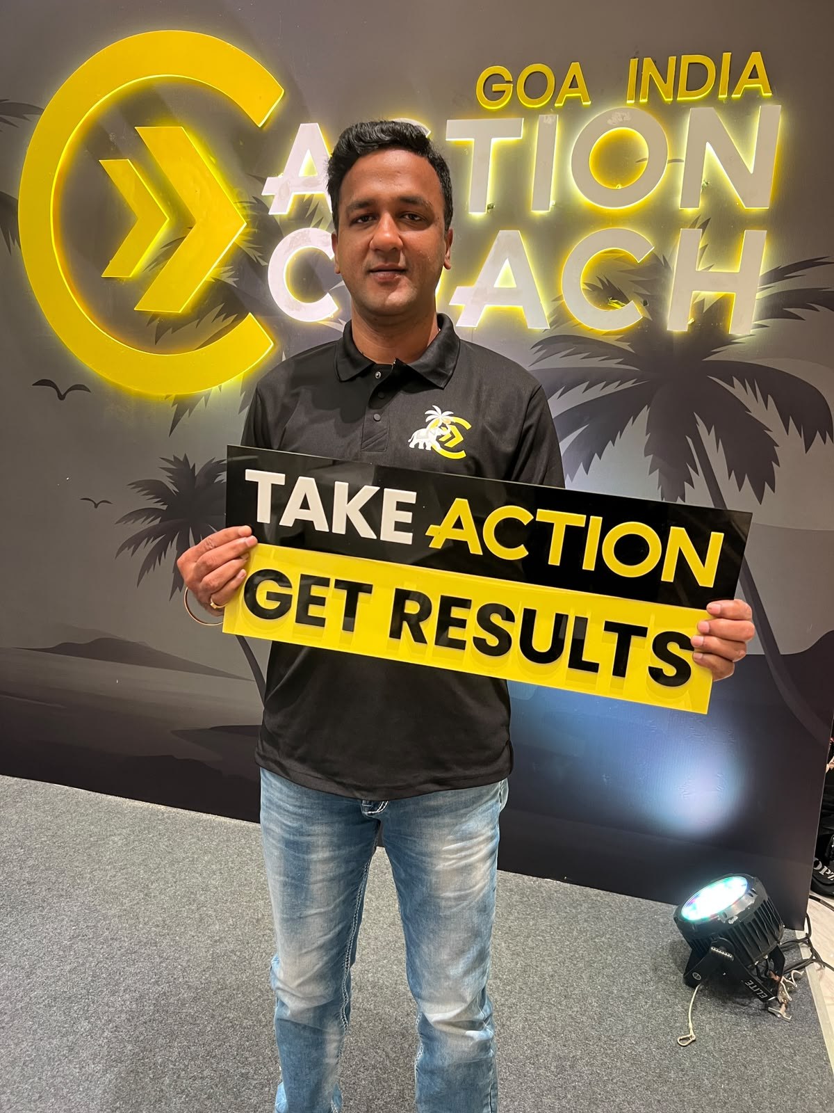 Coach Dhejo holding a Take Action Get Results sign at the ActionCOACH Global Conference in Goa